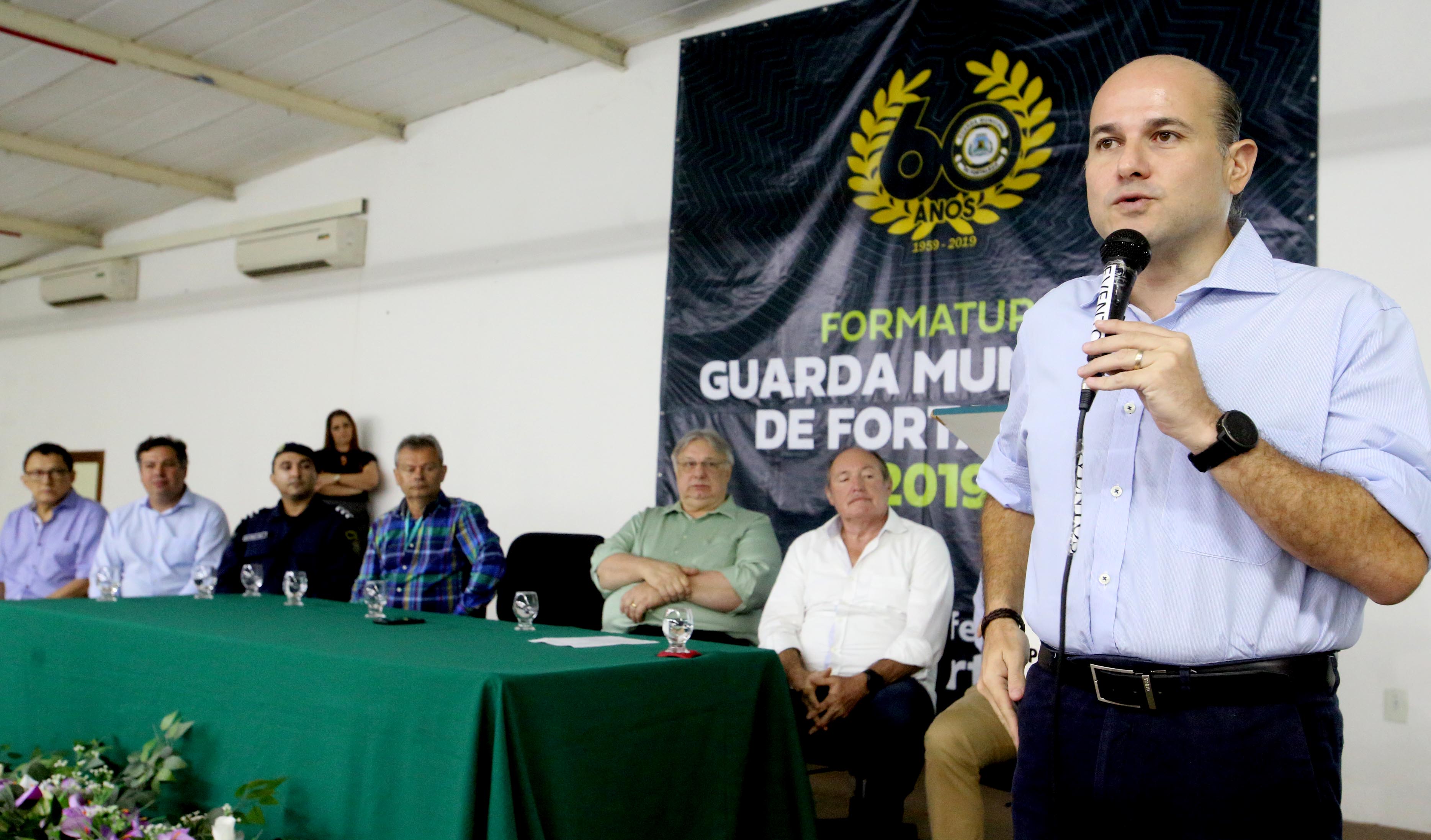 prefeito em primeiro plano falando o microfone e ao fundo secretários e vice-prefeito sentados assistindo